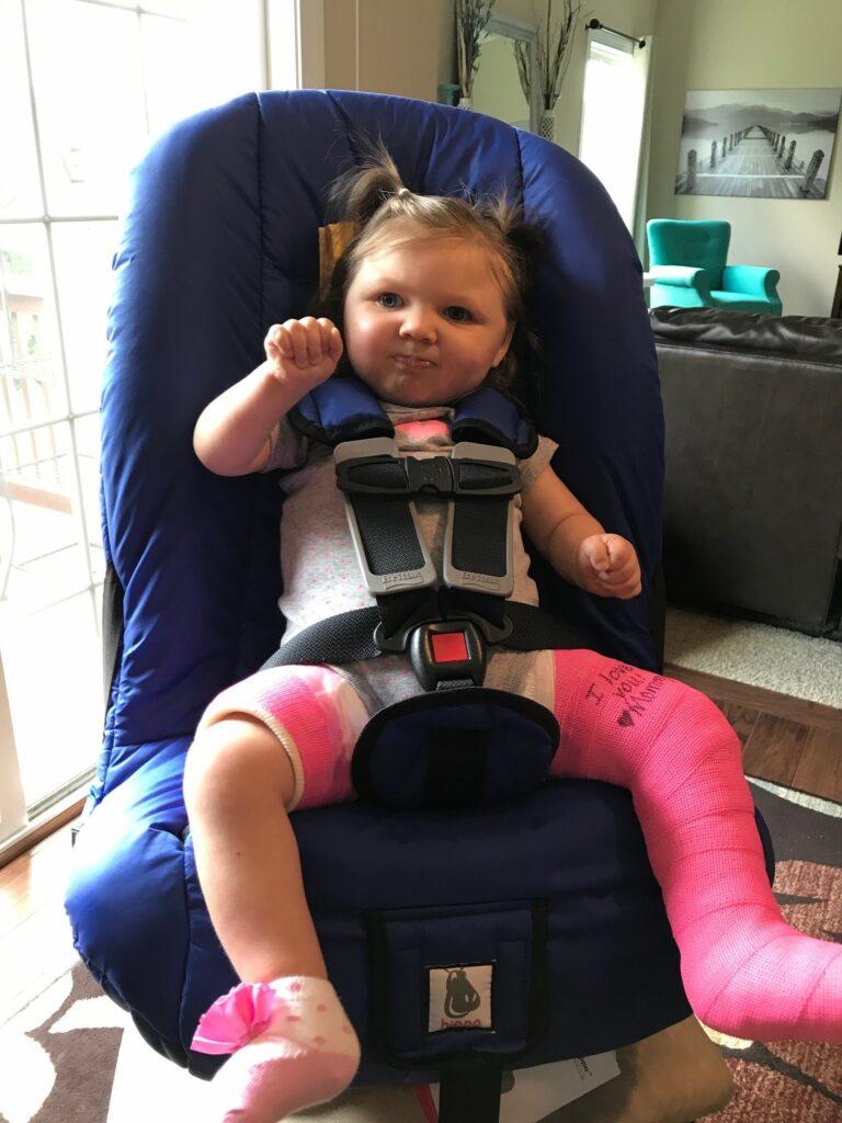 Elyse, then a toddler, sits in her adaptive car seat. She is wearing a hip spica cast.