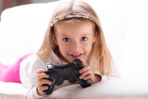 Adorable playful pretty little blond girl with a TV game controller lying on her bed grinning happily at the camera