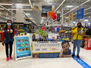Two people wearing face masks in a Walmart store standing on opposite ends of a table displaying a CMN banner.